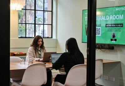 Glass-walled meeting room with two people working on laptops and a large green display screen.