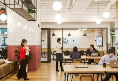 Bright coworking area with long wooden tables, tiled columns, and people working on laptops.