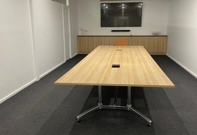 Meeting room with a long light-wood table, grey carpeting, and a wall-mounted screen.