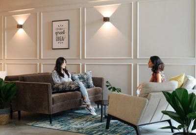 Lounge area featuring two women chatting on a brown leather sofa and cream armchair against a paneled wall.