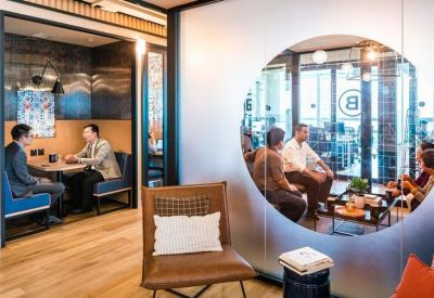 Lounge area with a circular glass partition looking into a bright meeting room with warm wood flooring.