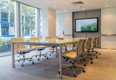 Bright conference room with a large glass table, tan leather chairs, and a wall-mounted monitor.