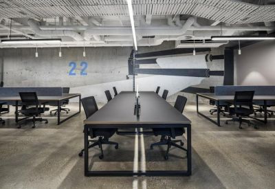 Large open-plan workspace with long black desks, ergonomic chairs, and industrial-style ceiling pipes.