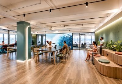 Bright open-plan communal area with a teal pillar, wooden seating, and a large ocean-themed digital screen.