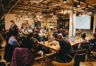 Large industrial-style event space with communal tables and people attending a presentation.