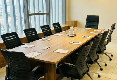 Long wooden boardroom table with eight black ergonomic chairs and artificial green window valance.