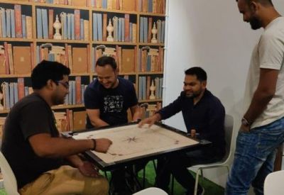 Group of colleagues playing carrom in a communal lounge with a library-themed feature wall.