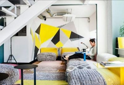 Creative breakout area under a staircase featuring a geometric yellow wall mural and floor cushions.