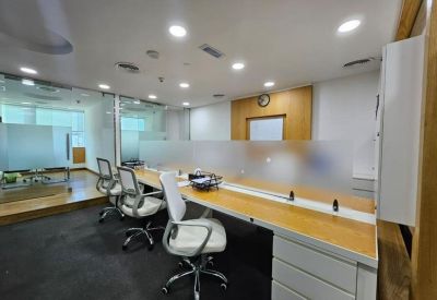 Bright open-plan workspace featuring long wooden desks, white office chairs, and frosted glass dividers.