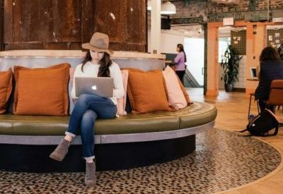 Woman working on a laptop on a curved green velvet banquette with orange pillows.