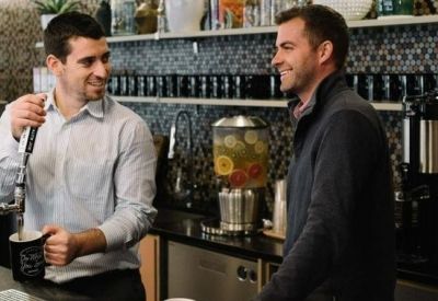 Two men talking in a modern office kitchen with coffee machines and a tiled backsplash.