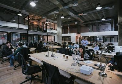 Industrial-style open-plan office with numerous desks, exposed piping, and high ceilings.