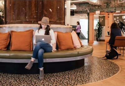 Person working on a laptop on a curved green leather bench with warm orange pillows.