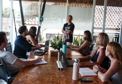 A facilitator leading a group discussion around a long wooden table in a bright meeting room.