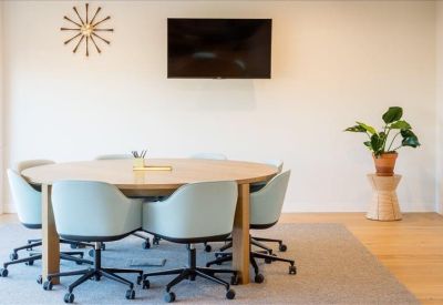 Minimalist circular meeting table with light blue ergonomic chairs and a wall-mounted screen.