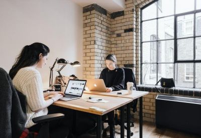 Two people working at wooden desks in a bright room with exposed brick walls and large windows.