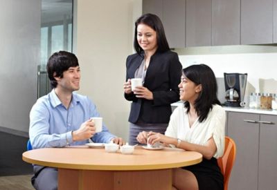 Three colleagues enjoying coffee and conversation in a modern office breakroom area.