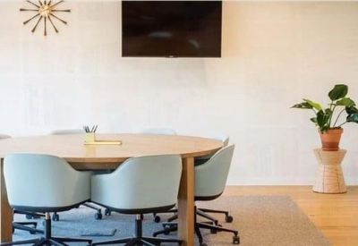 Bright meeting room with a round wooden table, light blue chairs, and a wall-mounted screen.