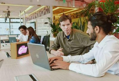 Open-plan office space where coworkers collaborate using laptops at a long desk.