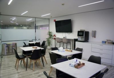 Modern breakroom with dining tables, coffee machine, and a wall-mounted television.