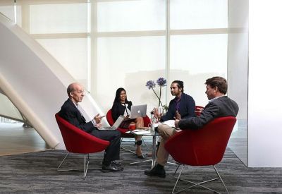 Informal lounge area with red bucket chairs and people working on laptops near large windows.