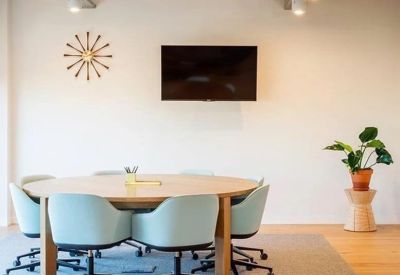 A round wooden meeting table surrounded by light blue chairs with a wall-mounted TV.