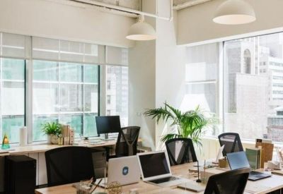Sunlit open-plan office with white desks, black chairs, and large windows overlooking the city.