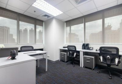 Bright corner office with three grey desks, ergonomic chairs, and large windows.