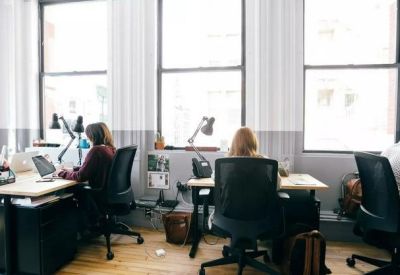 Open-plan office space with professionals working at rows of desks by large windows.