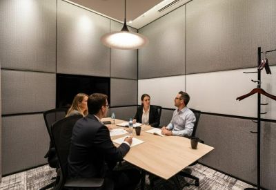 A modern meeting room with four people seated at a light wood table under a pendant light.