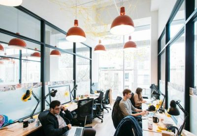 Sunlit open-plan office with multiple rows of desks, ergonomic chairs, and orange pendant lights.