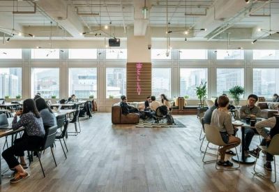 Open-plan office space with long communal desks, large windows, and people working in a bright environment.