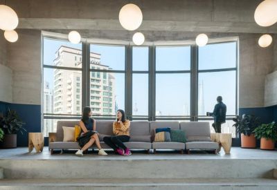 Bright breakout space with a long grey sofa, potted plants, and large windows overlooking city buildings.