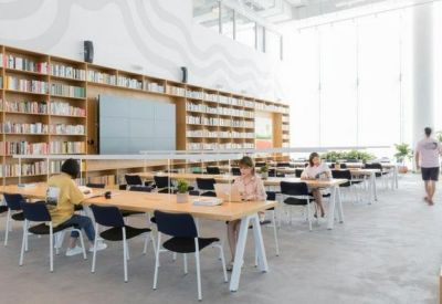 Open-plan office with long wooden desks, ergonomic chairs, and a large bookshelf backdrop.