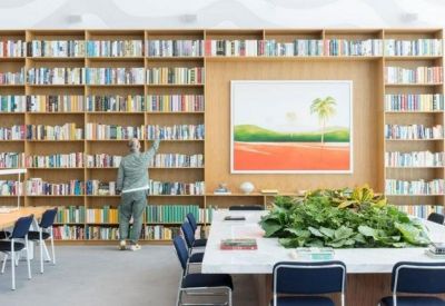 Bright library area featuring a large wooden bookshelf, a vibrant landscape painting, and a communal table with plants.