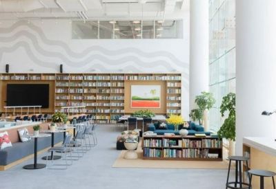 Wide-angle view of a sunlit coworking space with floor-to-ceiling windows, lounge seating, and wall-to-wall books.