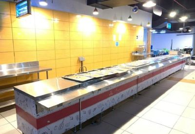 Long stainless steel cafeteria serving counter against a bright yellow tiled wall.
