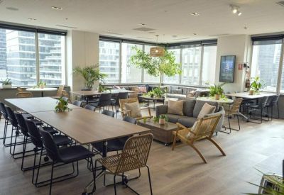 Sunlit open-plan office with long wooden tables and woven chairs near floor-to-ceiling windows.
