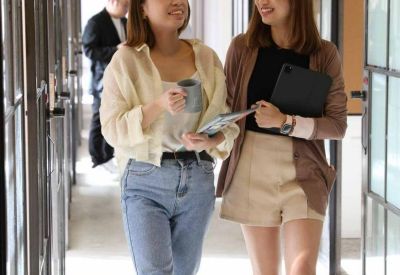 Two women walking through a bright corridor with glass-partitioned offices.