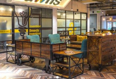 Lounge area featuring wooden seating with turquoise armchairs and glass-partitioned meeting rooms.