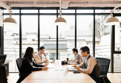 Bright office space with people working at a long wooden table against large windows.