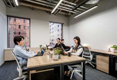 Modern open-plan office with people working at wooden desks under industrial ceilings.