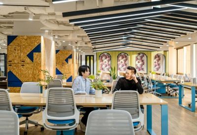 Coworking lounge with long wooden tables and vibrant wall art under a slatted ceiling.