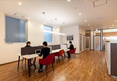 Open-plan office area with long white desks, red chairs, and large windows providing natural light.