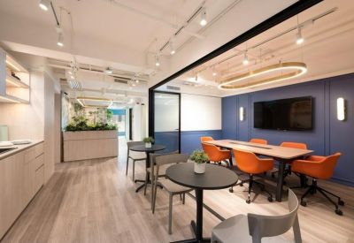 Spacious breakout area with orange chairs, meeting tables, and a blue accent wall with a large TV.