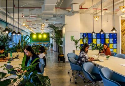 Open-plan office with people working at long tables under industrial pendant lighting.