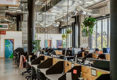 Large open-plan office space with rows of desks, hanging plants, and industrial ventilation.