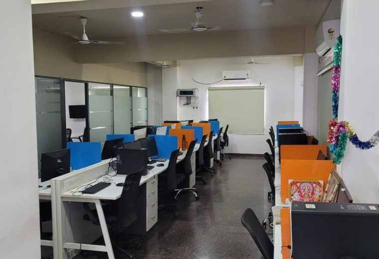 Open-plan workspace with rows of desks, blue and orange partitions, and ceiling fans.