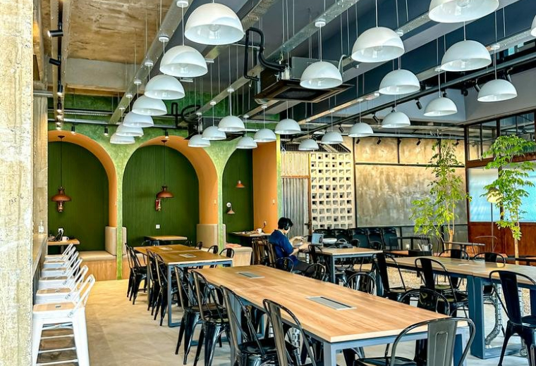 Industrial style cafe and communal area with long wooden tables and dome pendant lights.