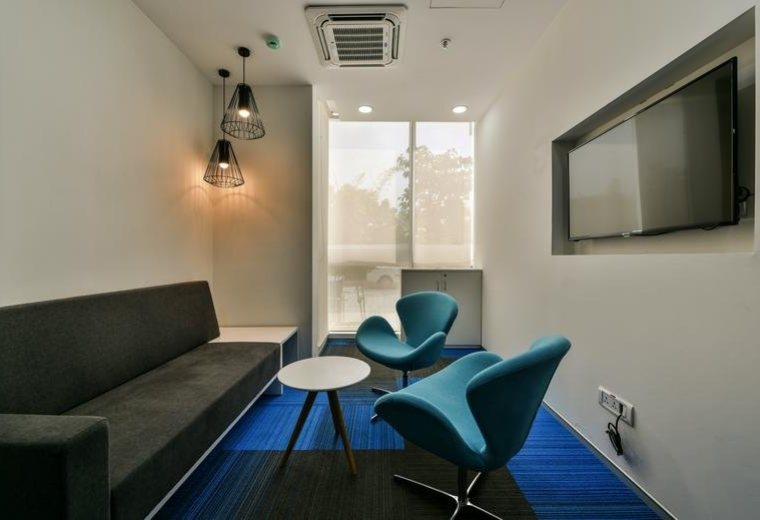 Meeting room at Ambience Mall, 7th Floor, NH 8 with a grey sofa, blue chairs, and wall-mounted TV.